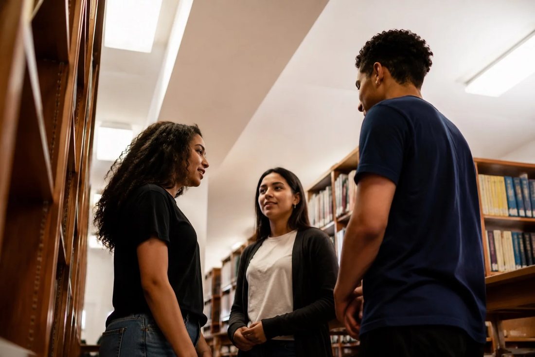 Alunos conversando em pé no corredor entre as prateleiras de uma biblioteca